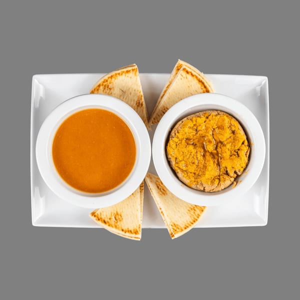 Two white bowls, one with orange soup and one with yellow dip, surrounded by pita bread.
